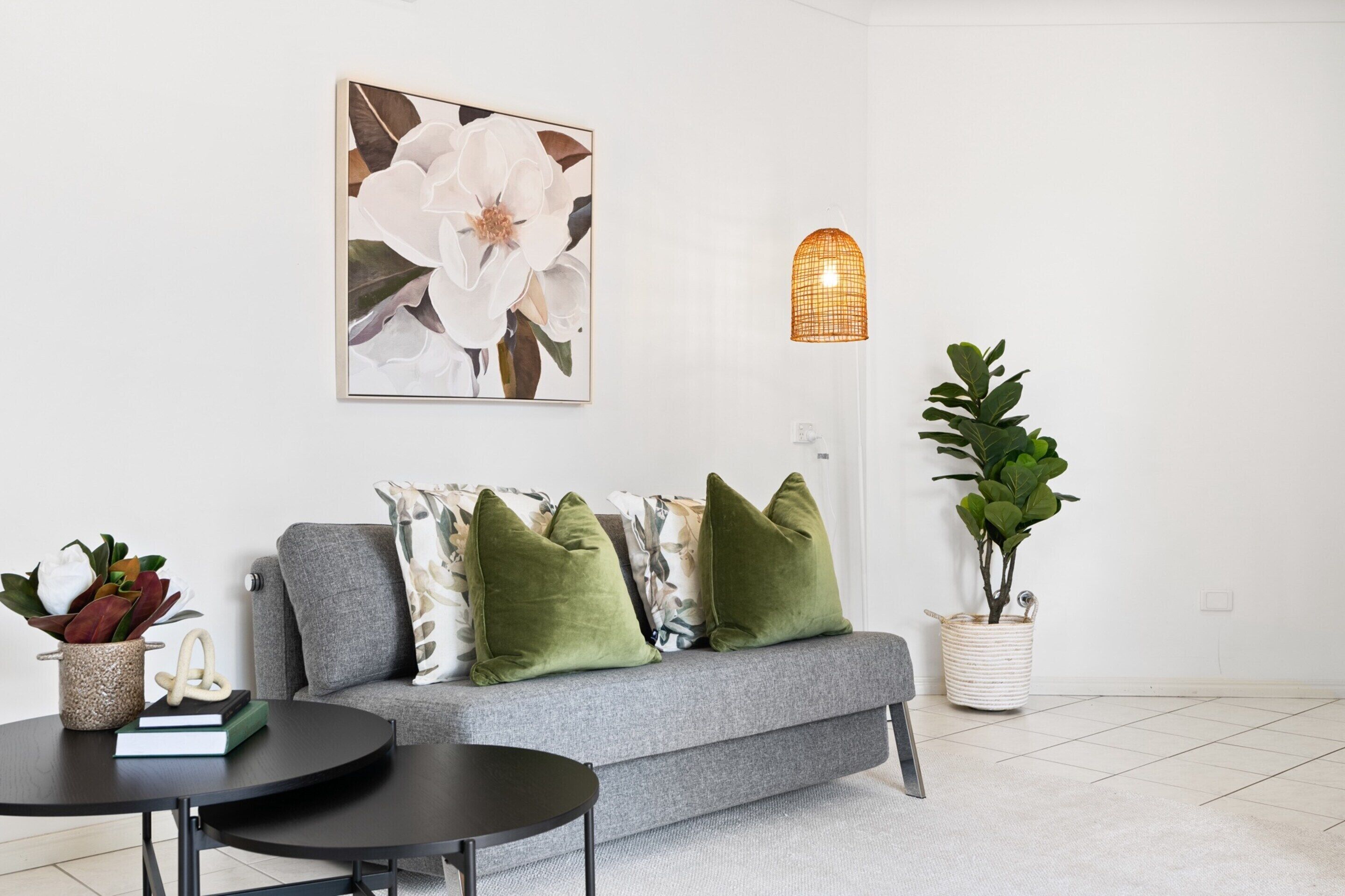 A modern living room with a grey sofa, green cushions, a round black table, a large floral painting, a woven pendant light, and a potted plant.