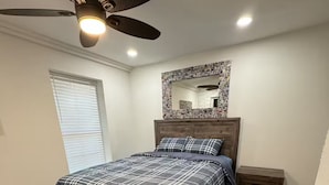 Ein Schlafzimmer mit Bett, Deckenventilator, Nachttisch, Spiegel und Fenster mit Jalousien.