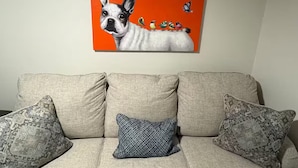 Ein beiges Sofa mit gemusterten Kissen und einem Wandbild, das einen Hund mit Vögeln zeigt.