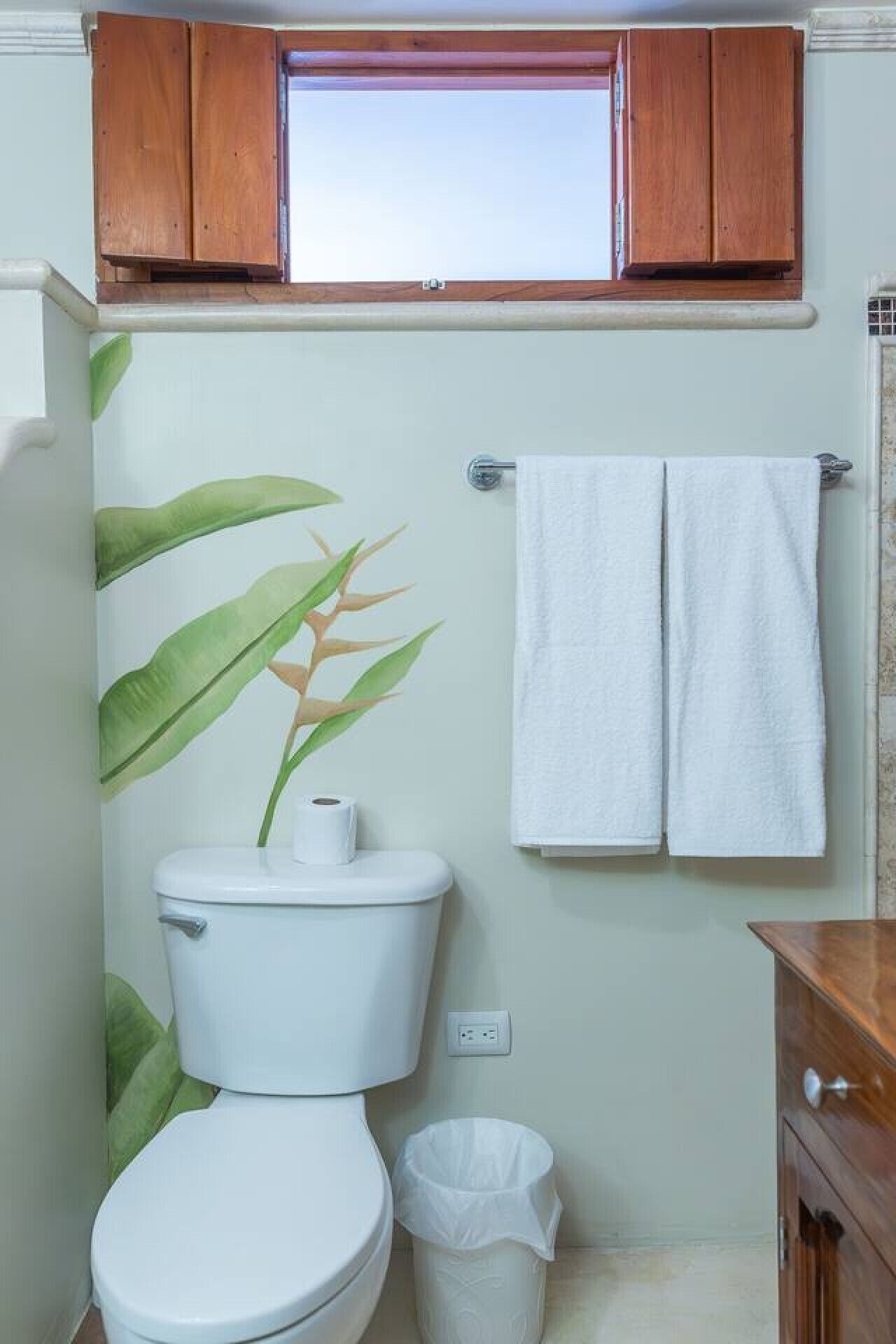 Un baño con un inodoro, un rollo de papel higiénico, un cubo de basura y una toalla colgada en un perchero.