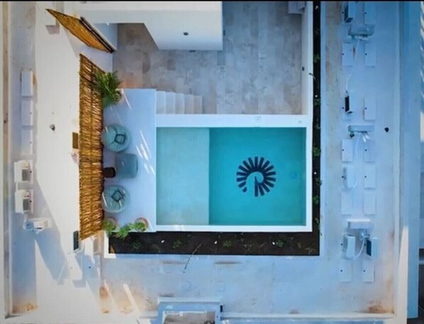 Vista aerea di una zona piscina con un logo a forma di sole, sedie a sdraio e un'area relax con tetto di paglia.