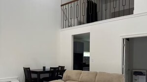 Un saló modern amb un sofà beix, un otomà estampat, una taula de dinar amb cadires, un ventilador de sostre i una escala que porta a un nivell superior.