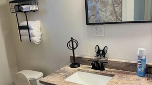 A bathroom with a white toilet, a sink with a granite countertop, a black towel rack, a mirror, and a bottle of hand soap.