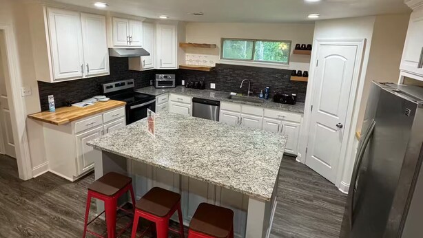 A modern kitchen with white cabinetry, a granite countertop island with red bar stools, stainless steel appliances, and a window providing natural light. - Frenchie Baker (Baker)