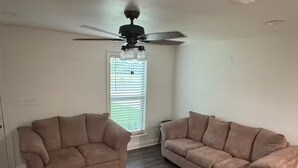 A room with two beige sofas, a ceiling fan, and a window with blinds. - Frenchie Baker (Baker)