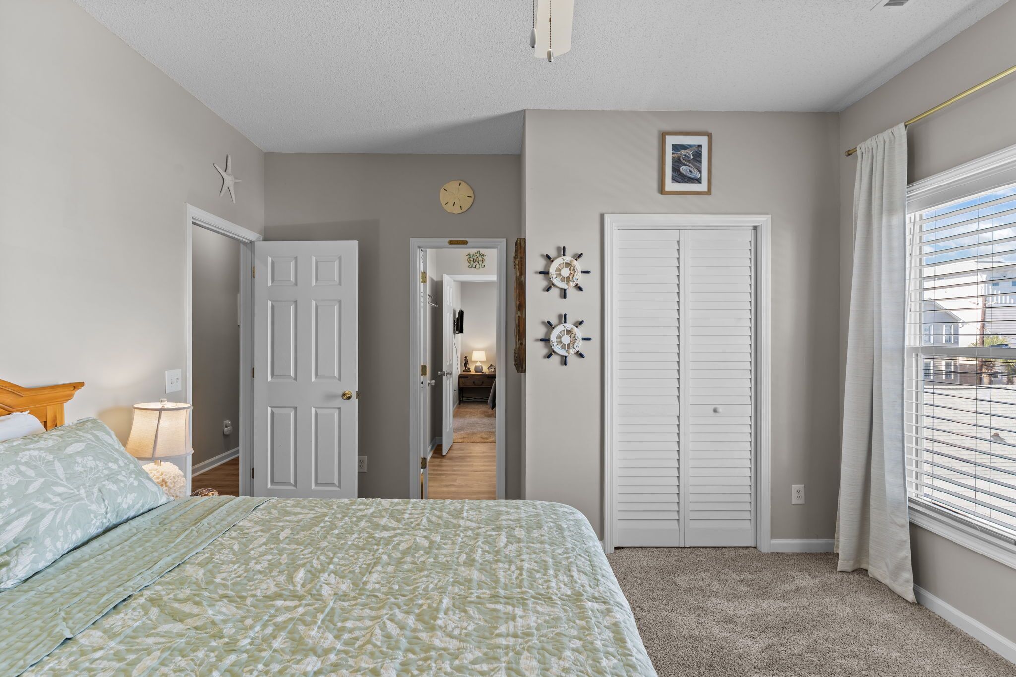A bedroom with a bed, a door, a clock, a window with blinds, and a hallway leading to another room.