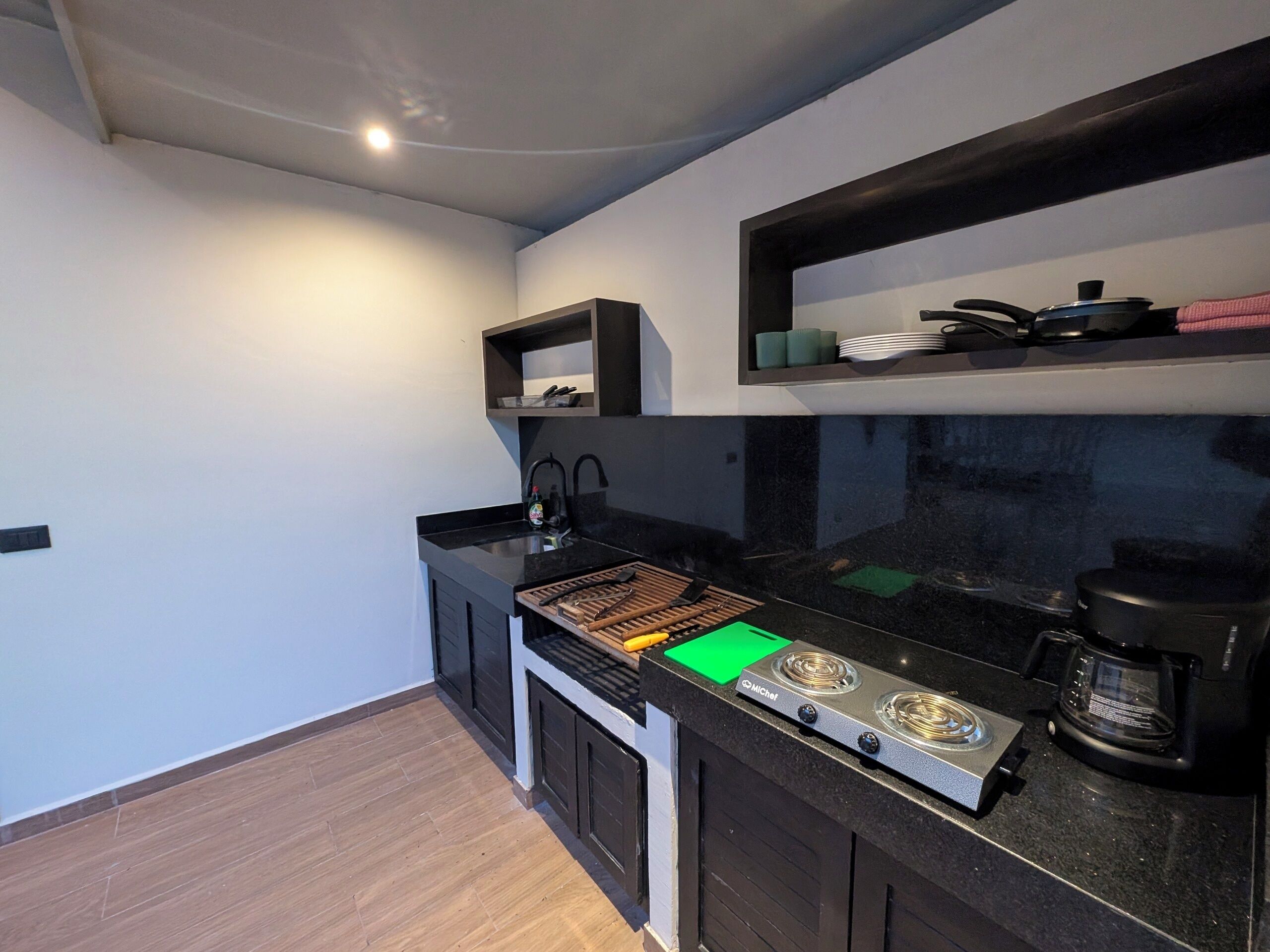A compact kitchen with a black countertop, a stove, a coffee maker, and a small shelf above the sink.