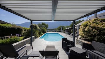 A covered outdoor area with a pool, lounge chairs, and a view of mountains.