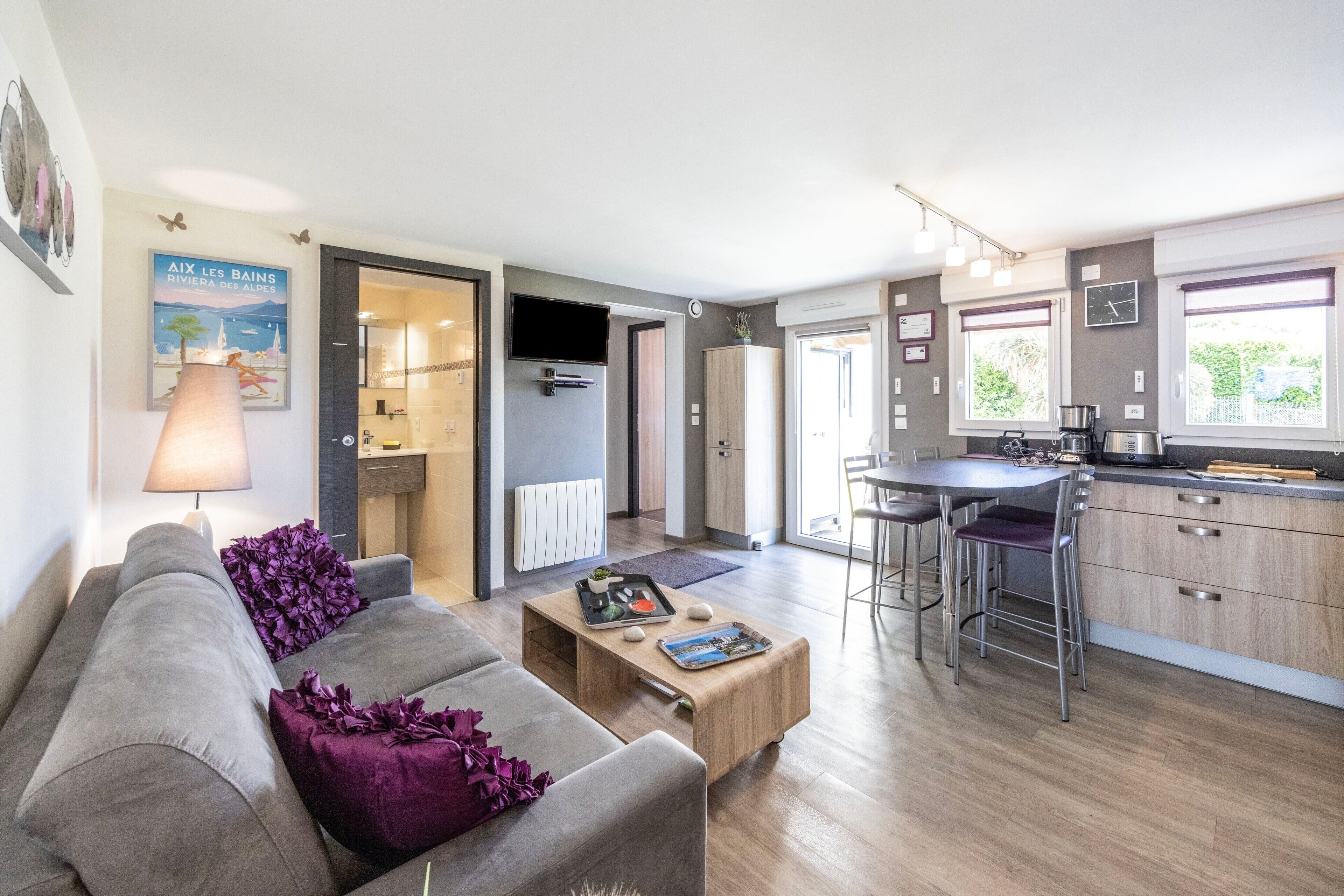 A modern living room with a grey sofa, a wooden coffee table, and a kitchen area with a bar stool.