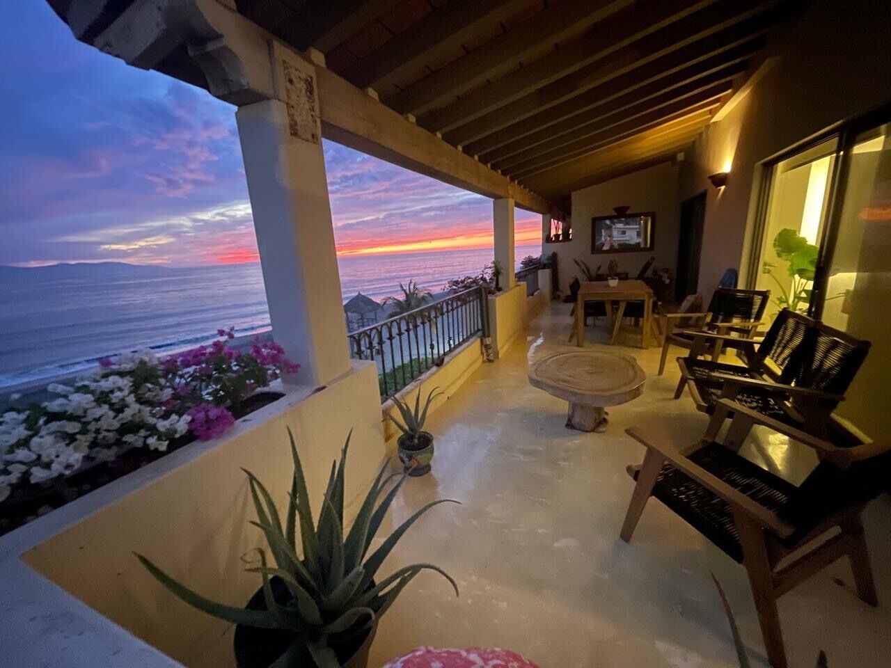 A balcony with a view of the sunset over the ocean, wooden furniture, and potted plants.