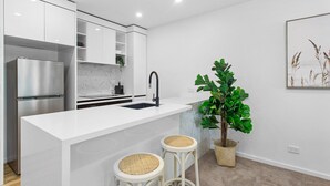 Una cuina moderna amb armaris blancs, un refrigerador d'acer inoxidable, un respent d'àmfores de marbre, un grifo negre i dos tamborets de vimet.