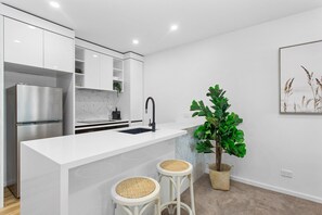 Eine moderne Küche mit weißen Schränken, einem Edelstahlkühlschrank, einer Marmor-Spritzschutzwand, einem schwarzen Wasserhahn und zwei Korbstühlen.