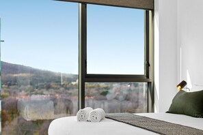 A hotel room with a large window, a bed with white linens, and a view of a hilly landscape. - Central 2-bed With Gym, BBQ & Stunning Views (Canberra)
