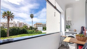 A balcony with a view of palm trees and buildings, a small table with a basket of flowers, and a washing machine. - Apartment 'Machadinho' with Private Pool, Wi-Fi and Air Conditioning (Albufeira)