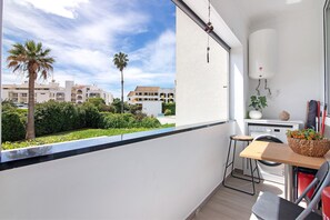 A balcony with a view of palm trees and buildings, a small table with a basket of flowers, and a washing machine. - Apartment 'Machadinho' with Private Pool, Wi-Fi and Air Conditioning (Albufeira)