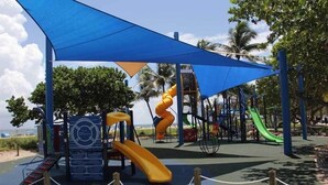 Ein Spielplatz mit Rutschen und Klettergerüsten, umgeben von Palmen und einem blauen Baldachin.