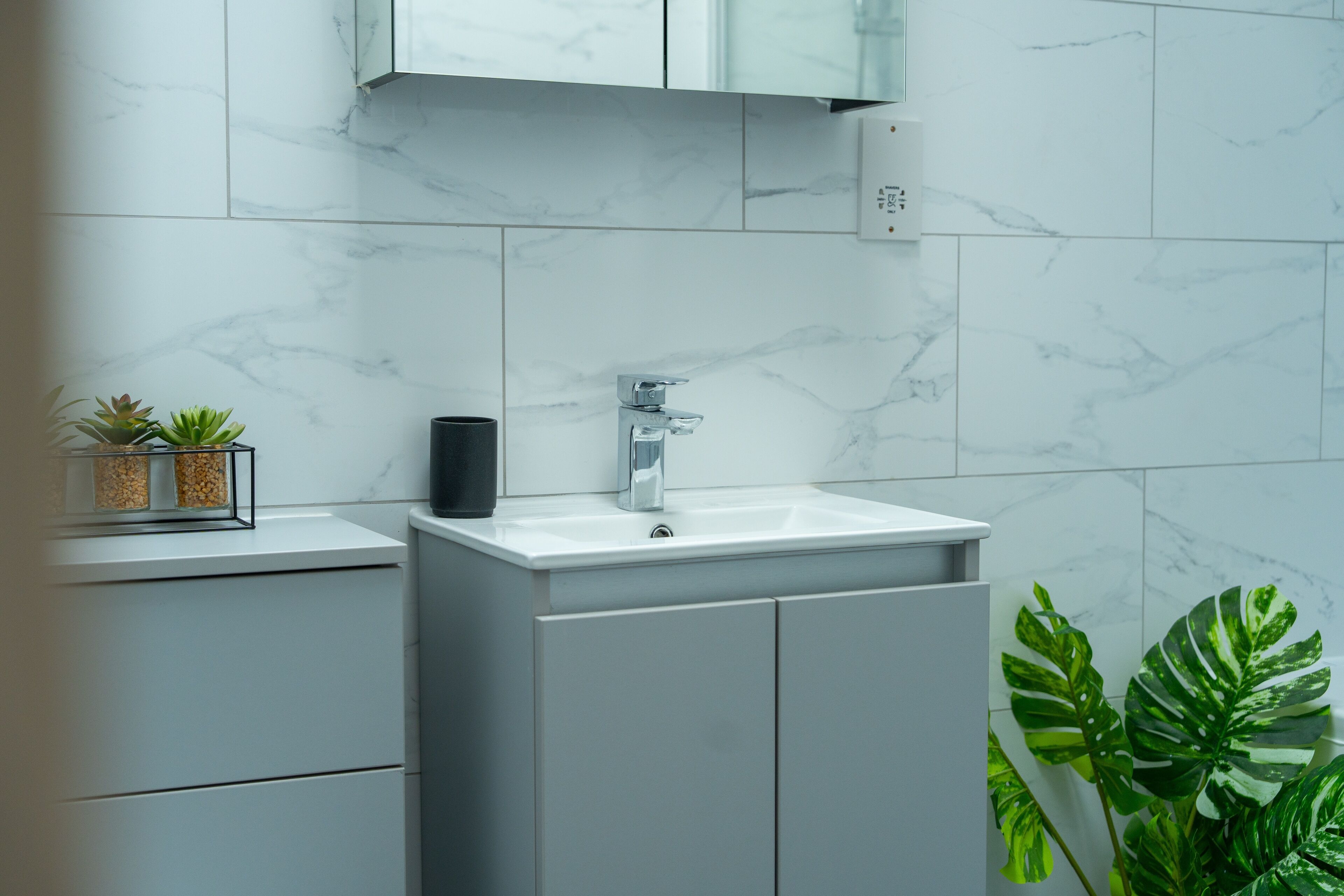 Un baño moderno con un lavabo de mármol blanco, armarios grises y una planta pequeña en la encimera.