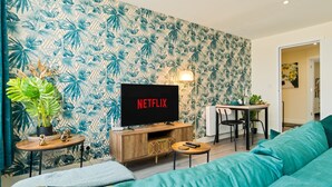 A living room with a television displaying 'NETFLIX', a wooden coffee table, a teal sofa, a dining area with a table and chairs, and a wall with a tropical leaf pattern. - Stylish City Centre getaway w/ Cathedral Views (Kent)