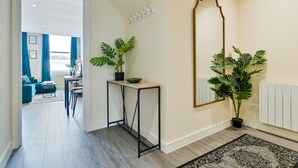 A modern hallway with a wooden floor, a black metal console table with a potted plant, a large mirror, and a view into a living area with blue curtains and a sofa. - Stylish City Centre getaway w/ Cathedral Views (Kent)
