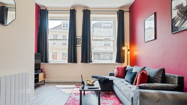 Un salon moderne avec un canapé gris, un tapis à motifs rouges et noirs, et une table basse sur laquelle se trouvent un croissant et une tasse.