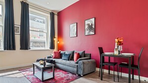 Un salon moderne avec un canapé gris, des coussins rouges, une table basse noire et un tapis rouge et noir. On y trouve une table à manger avec des chaises et un vase de fleurs. Un mur d’accent rouge est agrémenté de deux tableaux encadrés.