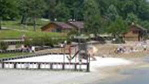 Zona junto a un lago con un muelle de madera que se adentra en el agua, rodeada de vegetación y algunos edificios.