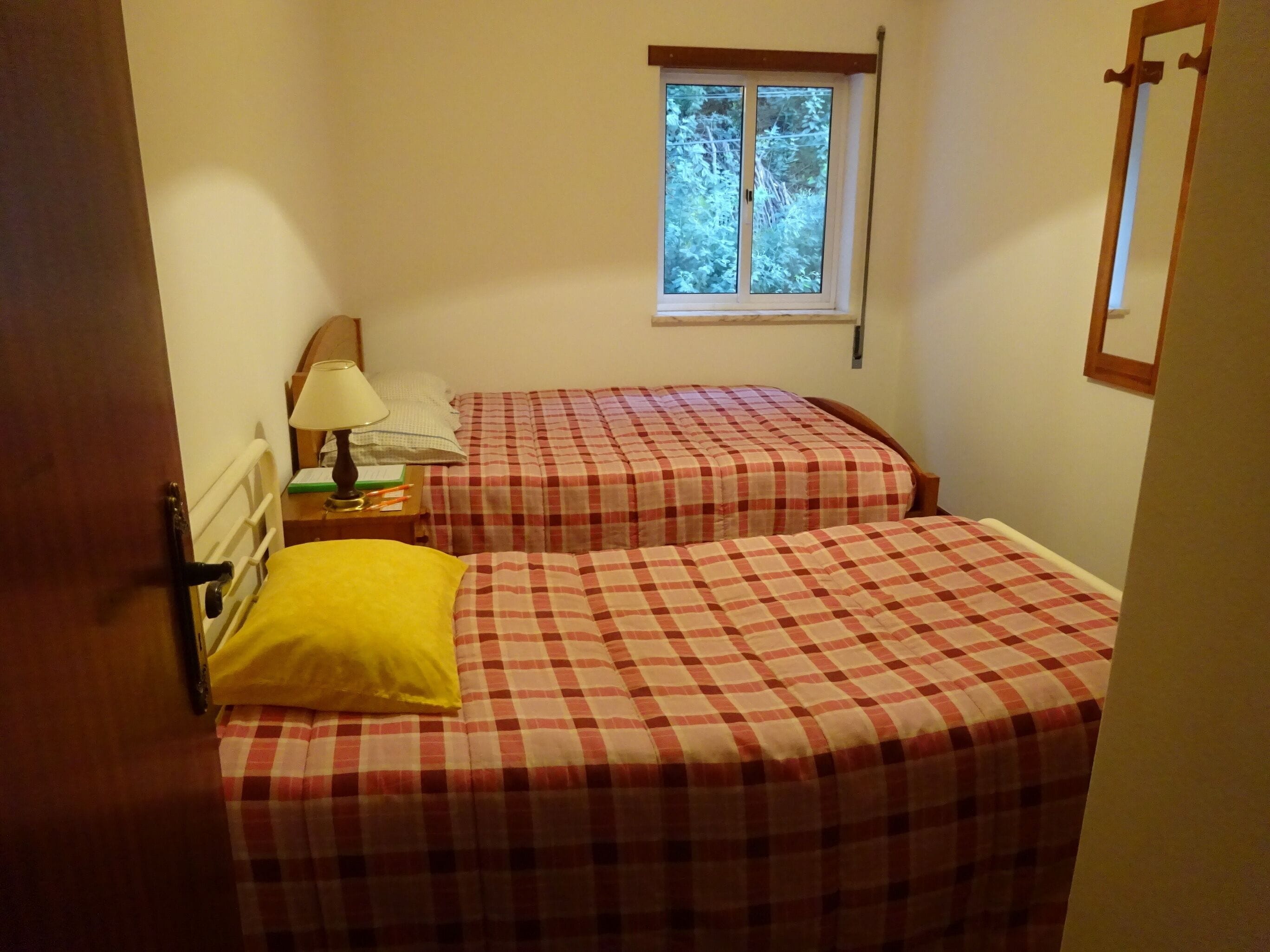 Un dormitorio con dos camas, cada una con colcha a cuadros, una mesita de noche con lámpara y una ventana con vista a árboles.