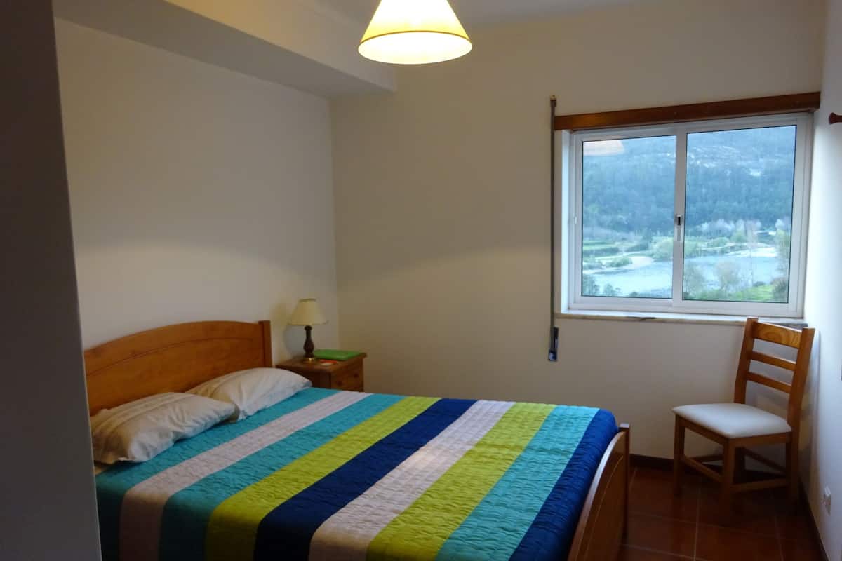 A bedroom with a bed, a chair, a nightstand, a lamp, and a window with a view of a landscape.