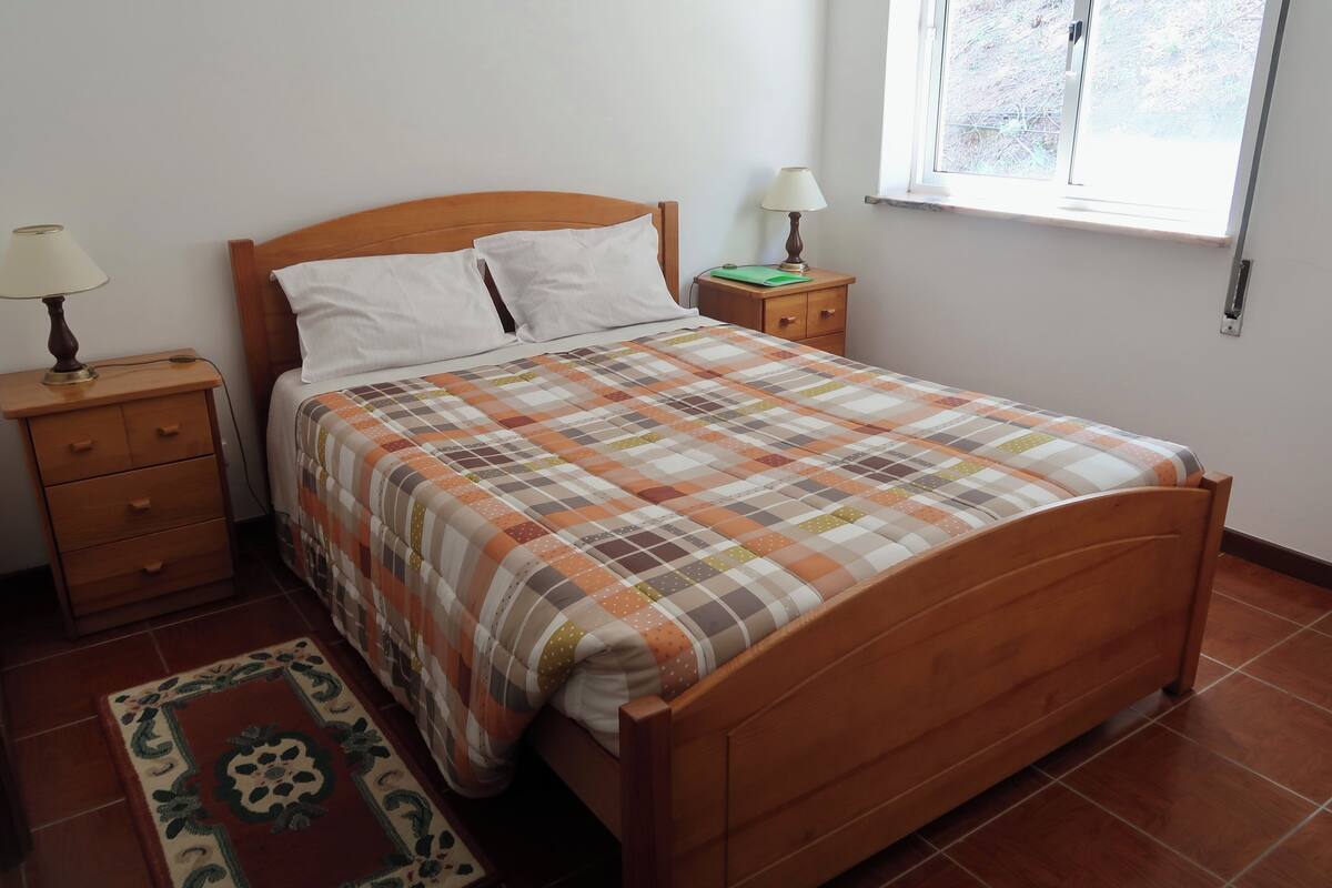 A neatly made bed with a checkered bedspread, two bedside tables with lamps, a small rug, and a window with a view of the outdoors.