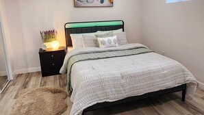Een slaapkamer met een bed, nachtkastje, wanddecoratie, raam en ventilatie rooster in het plafond.