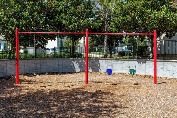 A playground with a red swing set and a blue bucket swing, surrounded by trees and a brick wall. - 3BR Disney Townhome with Private Pool – Walk to Windsor Hills Water Park (Kissimmee)
