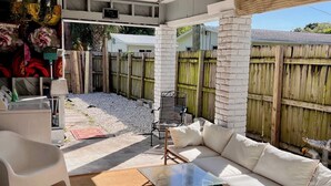 Eine sonnendurchflutete Terrasse mit einem weißen Sofa, einem Glastisch und einem Stuhl. Im Hintergrund sind ein Holenzaun und eine Backsteinmauer zu sehen.