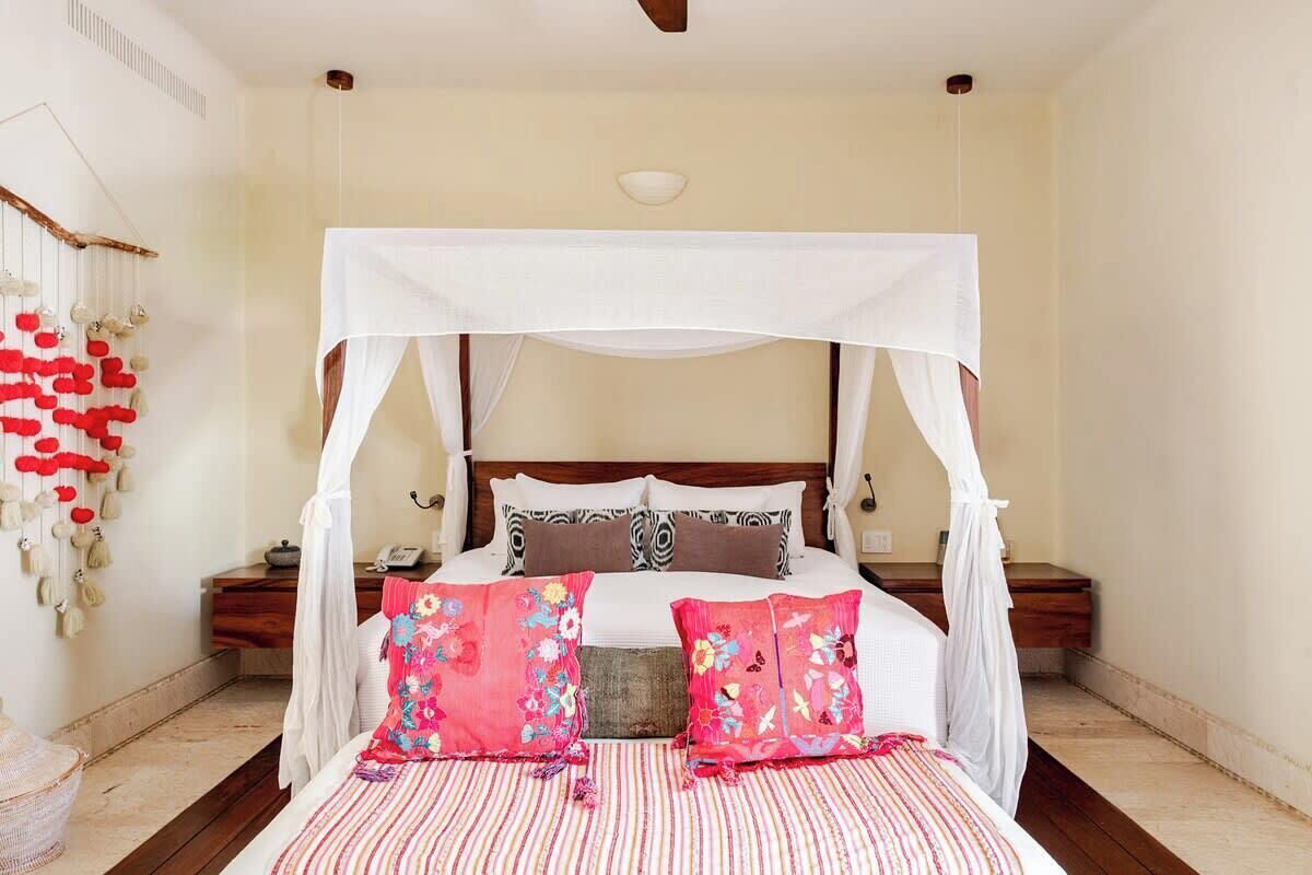 Una cama con dosel, almohadones florales y cubrecama a rayas en una habitación con suelo de madera y pared blanca.