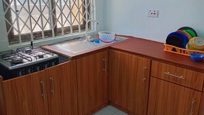 A kitchen with wooden cabinets, a gas stove, a window with a metal screen, and a sink with a dish rack. - Home Stay In Accra, Travellers With Kids Are Welcomed. 5 mins Walk Away To Beach (Accra)