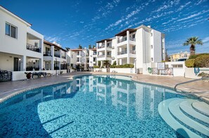 Una piscina rodeada de edificios blancos con balcones, cielo azul despejado y palmeras.