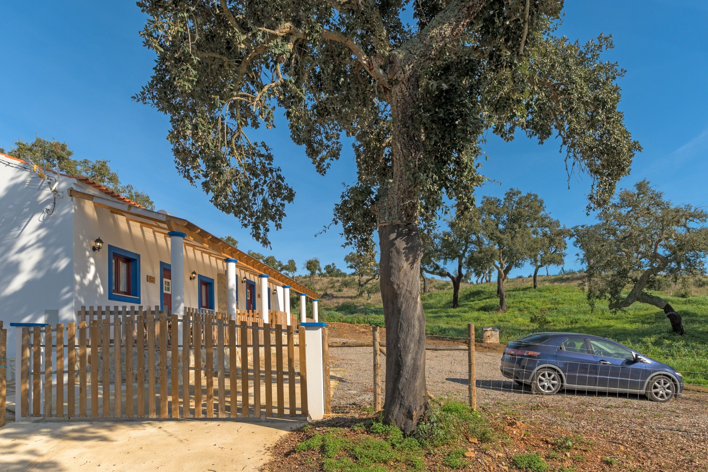 Un edificio bianco con una recinzione di legno, un'auto parcheggiata e un albero grande in un ambiente rurale.