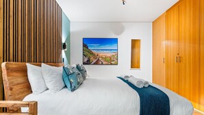 A bedroom with a wooden headboard, a large bed with white linens, a blue and white pillow set, a blue throw blanket, and a painting of a beach scene on the wall. - Greenmarket Place 503 (Cape Town)