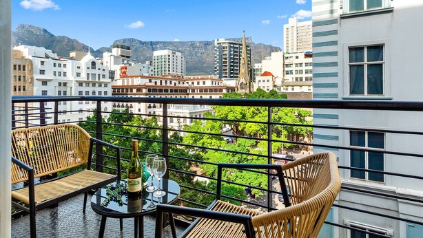 A balcony with wicker chairs and a table, overlooking a cityscape with buildings and a mountain in the background. - Greenmarket Place 503 (Cape Town)