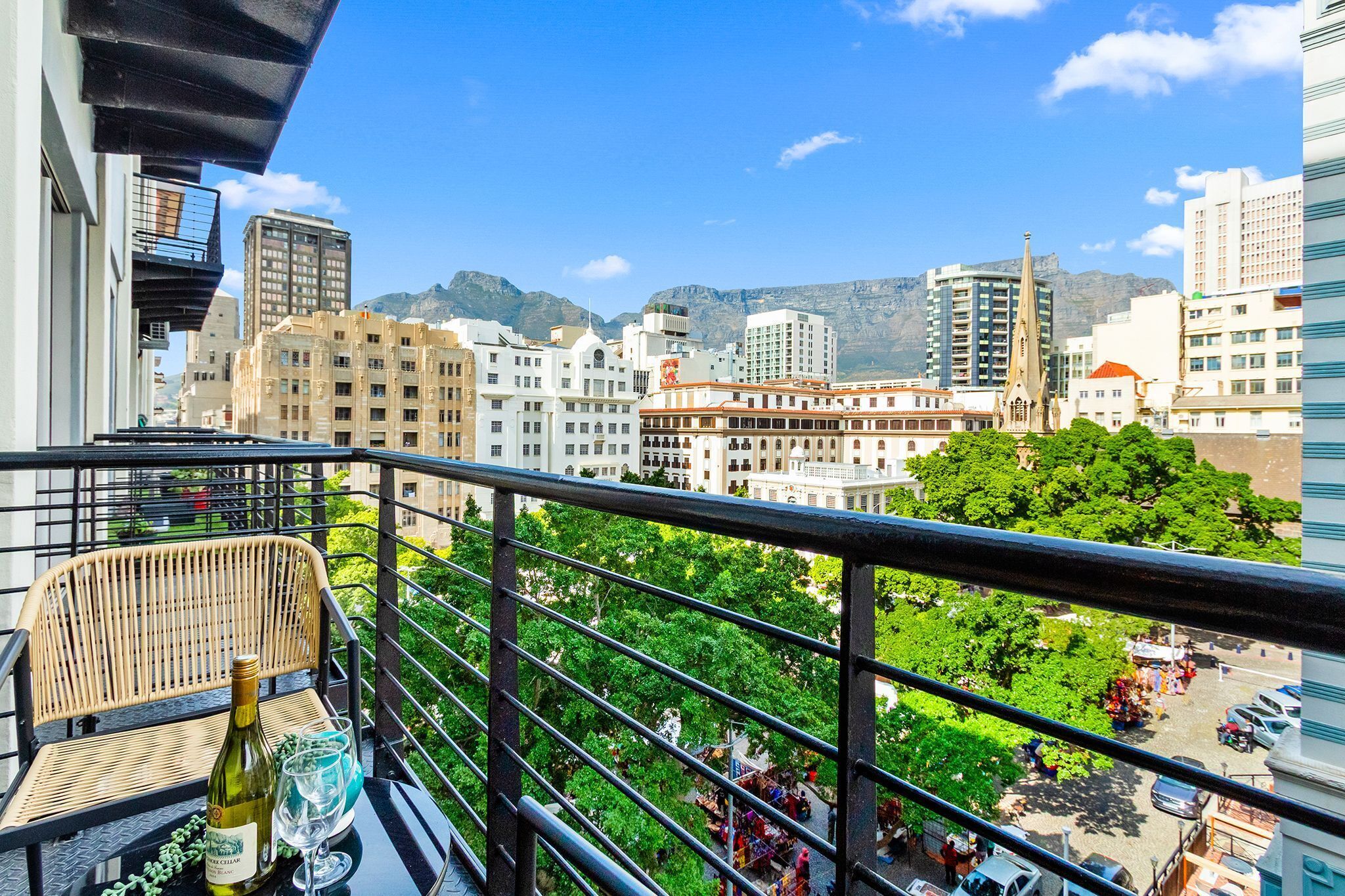 Varanda com cadeira de vime, uma garrafa e copos, com vista para um horizonte urbano com edifícios e árvores.