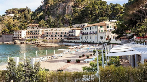 Un hôtel situé sur la côte, composé de plusieurs bâtiments, avec une plage équipée de transats et une falaise rocheuse en arrière-plan.