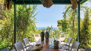 Une table en bois, dressée pour le dîner, avec des chaises, un bol de noix, une bouteille de vin et des verres, sur une terrasse offrant une vue sur l’océan.