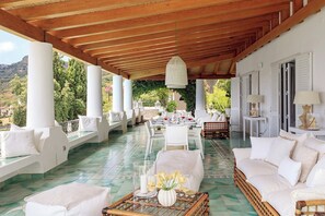 A covered outdoor seating area with white furniture, a dining table set with red wine glasses, and a view of greenery and hills in the background.