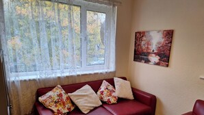 Un sofá de cuero rojo con cojines estampados, una ventana con cortinas de gasa y un cuadro de árboles otoñales en la pared.