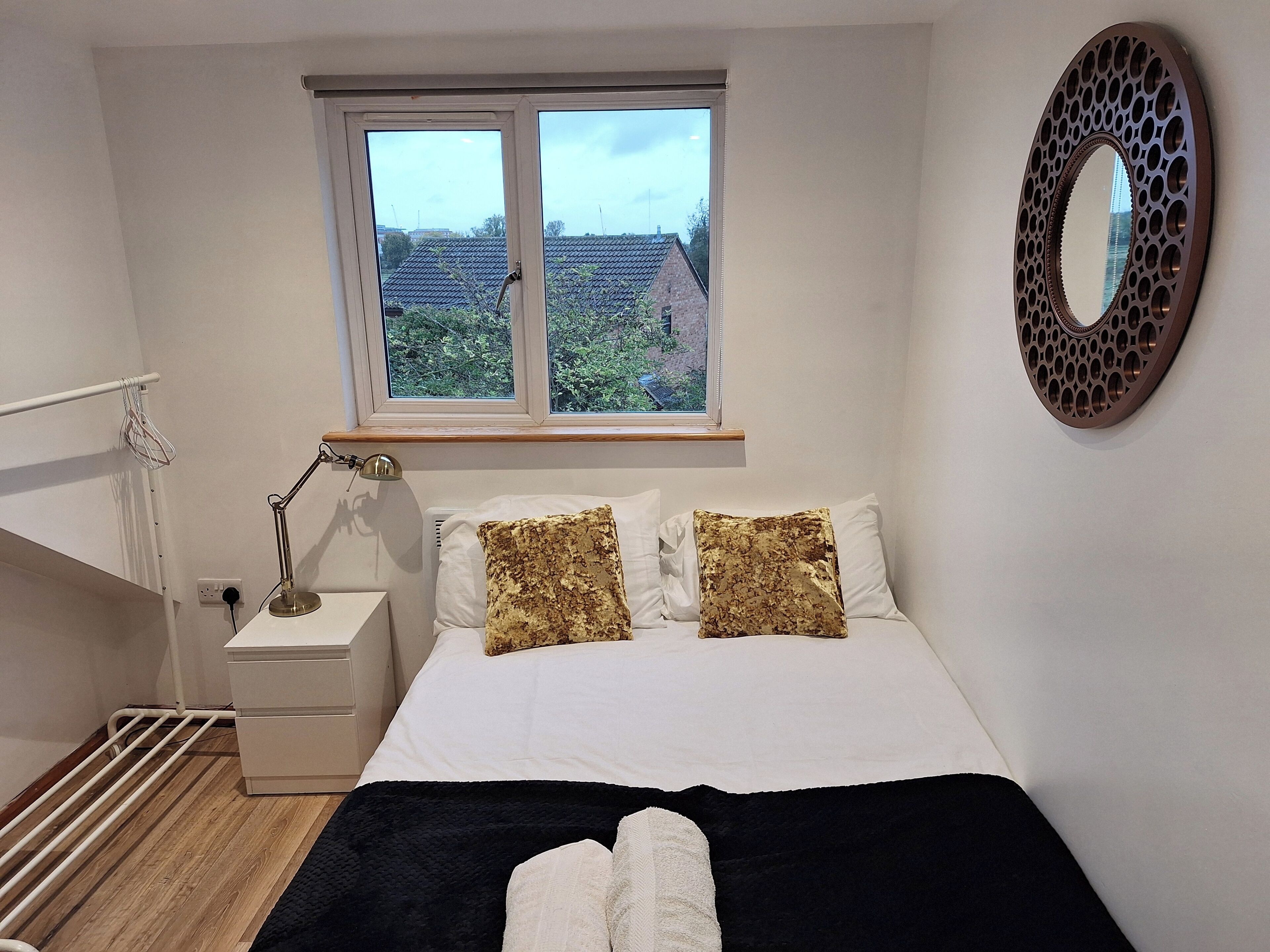 A bedroom with a bed, white pillows, a black blanket, a bedside lamp, a radiator, and a window with a view of buildings and trees.