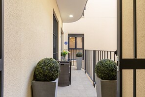 Um corredor estreito com varanda, plantas em vasos, uma mesa com cadeiras e uma porta com aviso.