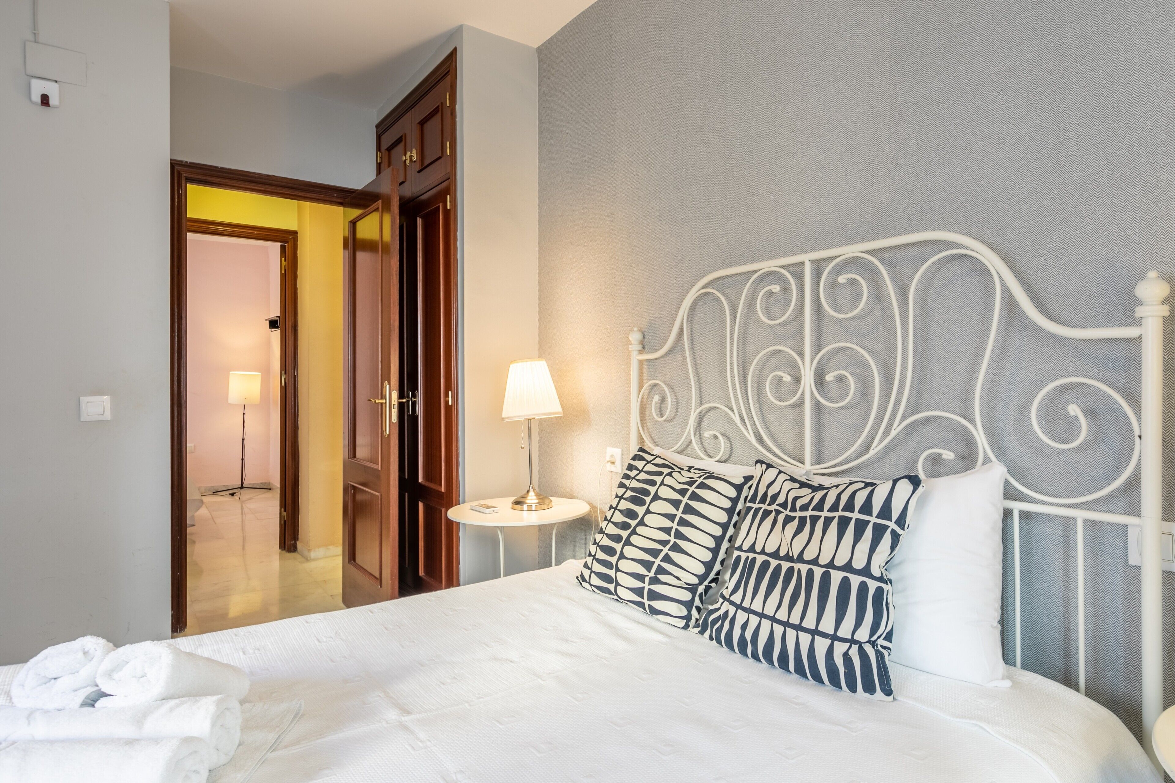 Una cama bien hecha con cabecera blanca, lámpara de noche y almohadillas estampadas.