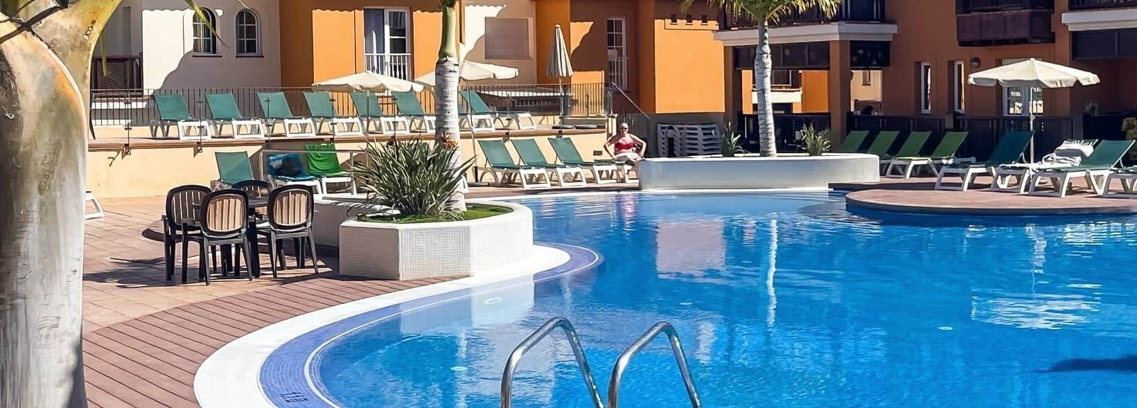Une piscine entourée de chaises longues et d’une table, le tout bordé d’un bâtiment doté de balcons.