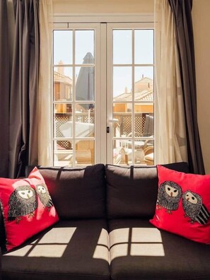 Ein Zimmer mit einem dunkelgrauen Sofa, zwei roten Kissen mit Eulenmotiven und Blick durch eine Glastür auf einen Außenbereich mit Möbeln.