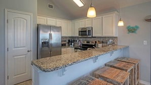 A kitchen with white cabinets, a stainless steel refrigerator, a microwave, a stove, granite countertops, and wooden stools. - Bright Coastal Escape w/ Games, Swing Chairs & Minutes to Port A Beach Captain Kirk by Avantstay (Port Aransas)