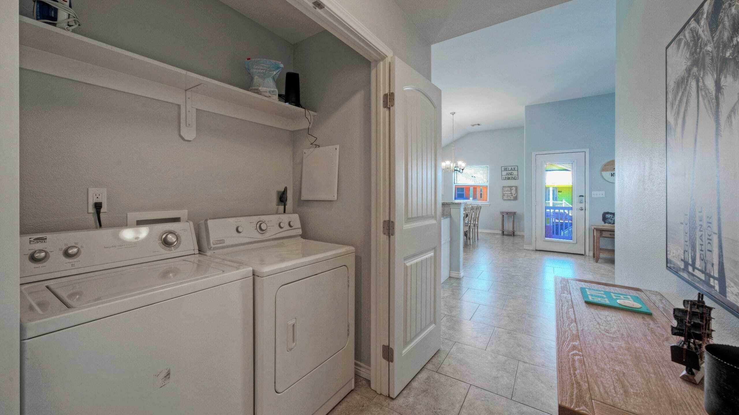 A laundry room with two front-loading washers and dryers, a shelf above the dryers, and a door leading to a dining area with a table and chairs.